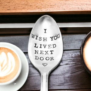 May include: A silver spoon with the stamped text "I WISH YOU LIVED NEXT DOOR" and a small heart symbol. The spoon is on a dark wooden surface, with a cup of coffee and a saucer in the background.