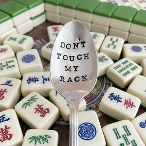 May include: A silver spoon with the text "DON'T TOUCH MY RACK" in black lettering. The spoon is surrounded by ivory-colored Mahjong tiles with green and red markings. Green-topped tiles are stacked in the background.