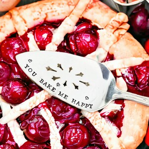 May include: A cherry pie with a decorative pie server. The pie server is silver and has the words "YOU BAKE ME HAPPY" stamped on it. The pie is filled with cherries and has a golden crust.