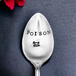 May include: A silver spoon with the word "POISON" stamped on the bowl in a bold, black font. Below the word is a small skull and crossbones symbol. The spoon is set against a dark background.