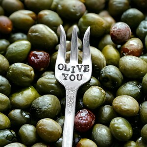 May include: A silver fork with the words "OLIVE YOU" stamped on it, surrounded by green olives. The fork has a floral design on the handle. Some olives are a reddish-brown color.