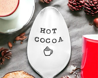 May include: A silver spoon with the words "HOT COCOA" and a graphic of a mug with whipped cream. The spoon is surrounded by a white teacup with cocoa, a red mug, pinecones, and other decorative elements on a gray surface.