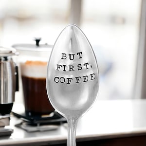 May include: A silver spoon with the text "BUT FIRST, COFFEE" stamped on the bowl. The spoon is in focus, with a blurred background showing a coffee drink and a coffee maker. A perfect gift for coffee lovers.