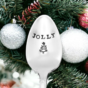 May include: A silver spoon with the word "JOLLY" stamped on the bowl, along with a small Christmas tree design. The spoon is set against a backdrop of Christmas tree branches and ornaments.