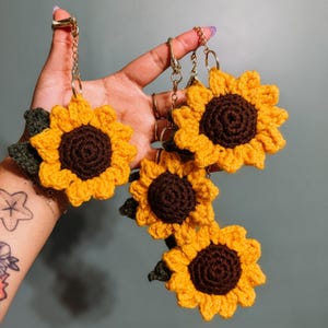 May include: A set of three sunflower keychains made from crocheted yarn. Each sunflower features a dark brown center, bright yellow petals, and a small dark green leaf attached to the side. Each flower is connected to a silver metal chain and ring.