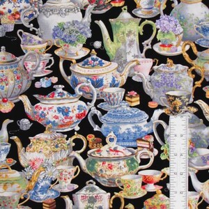 May include: A black fabric featuring a repeating pattern of various colorful teapots, teacups, and small cakes. A white L-shaped ruler is placed over the fabric, showing measurements in inches along the bottom and right edges of the image.