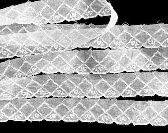 May include: A length of white lace trim featuring a repeating diamond pattern with small embroidered rose motifs centered within each diamond shape. The fabric has a semi-sheer texture with scalloped edges along the bottom border of the trim.