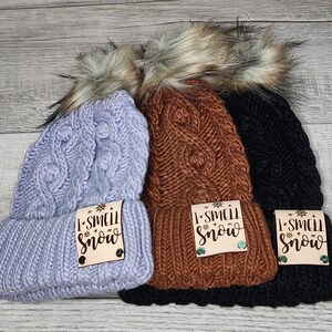 May include: Three knitted winter hats with faux fur pom poms. Each hat has a wooden tag that says "I Smell Snow". The hats are in lavender, brown, and black.