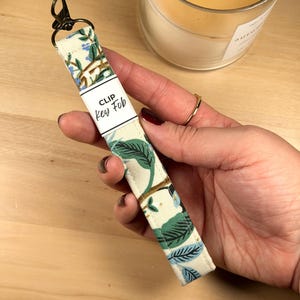 May include: A fabric key fob with a cream background featuring a botanical pattern of green leaves and small blue flowers. The strap includes a dark metal lobster clasp at the top and a white paper label printed with the text CLIP Key Fob.