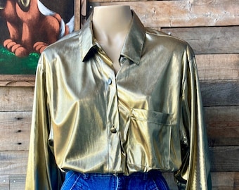 May include: A gold metallic button-up shirt with long sleeves and a single pocket on the left side. The shirt is worn over blue jeans.