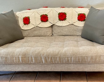 May include: A light beige sofa with a crocheted throw featuring red roses. The sofa has two gray throw pillows.