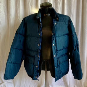 May include: A dark blue puffer jacket with a zipper and snap closure. The jacket has a quilted design and two front pockets.