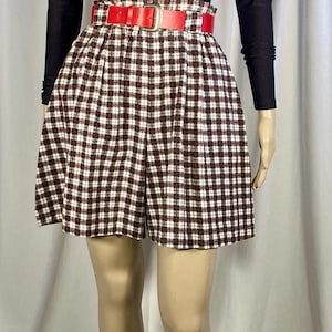 May include: A black and white plaid overall skirt with a red belt. The skirt has a high waist and is made of a thick fabric. The model is wearing a black long-sleeved shirt underneath.