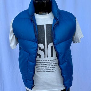 Peut inclure: Un gilet matelassé bleu vif avec une fermeture éclair complète, porté sur un t-shirt blanc. Le t-shirt présente un grand graphisme noir et blanc avec le mot "SO" et une liste de dates et de lieux. Le gilet est présenté sur un mannequin.
