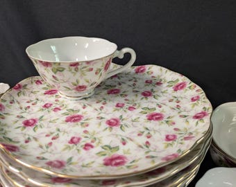 May include: A stack of white porcelain plates and a teacup with a floral rose pattern in pink and green. The teacup sits on a matching plate. The edges of the plates and teacup are trimmed with gold.