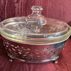 May include: A vintage serving dish with a clear glass lid and a silver-colored metal base. The lid features etched floral designs and a knob handle. The base has decorative cut-out patterns and rests on small feet. The dish is designed for serving food.