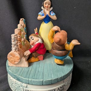 May include: A ceramic musical figurine featuring Snow White, two dwarfs, and a stack of forest animals on a round base. The base is painted with a wood-plank texture and features the text "SNOW WHITE AND THE SEVEN DWARFS" printed on the side.