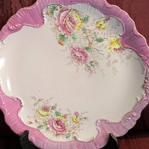 May include: An oval-shaped, antique serving platter with a pink scalloped edge. The center is white, adorned with pink and yellow roses and green leaves. The platter is supported by a black stand, showcasing a vintage aesthetic.