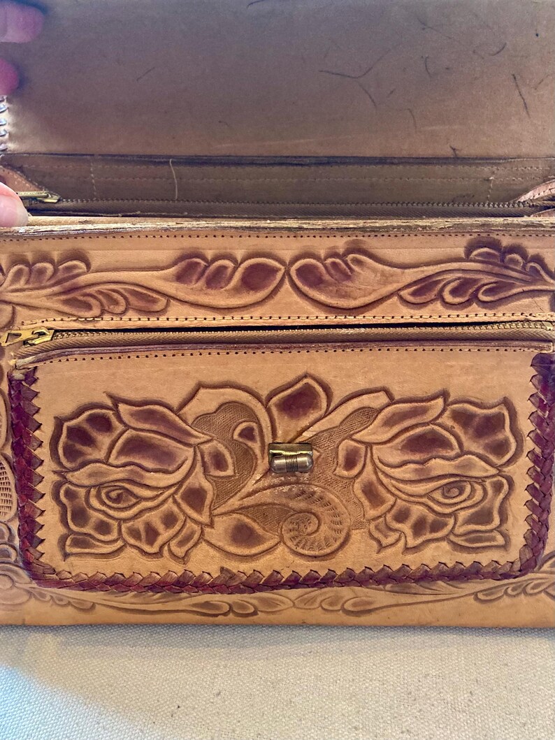 Pode incluir: Uma bolsa de couro castanho claro com um design floral. A bolsa apresenta um padr&atilde;o de rosas em relevo na frente, um bolso com z&iacute;per e um fecho de metal. As bordas s&atilde;o costuradas com linha castanha mais escura.