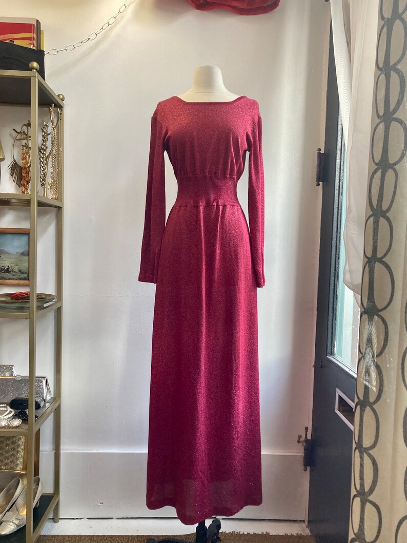 May include: A long, burgundy knit dress with a fitted bodice and a flowing skirt. The dress has a subtle shimmer and a slightly stretchy fabric.