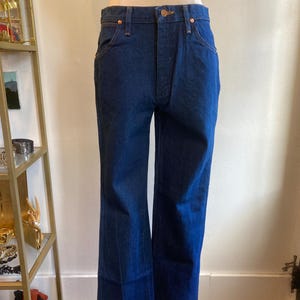 May include: Dark blue denim jeans with a classic five-pocket design and a slight flare at the hem. The jeans are displayed on a mannequin, showcasing the fit and style. The jeans have copper-colored rivets and a button closure.