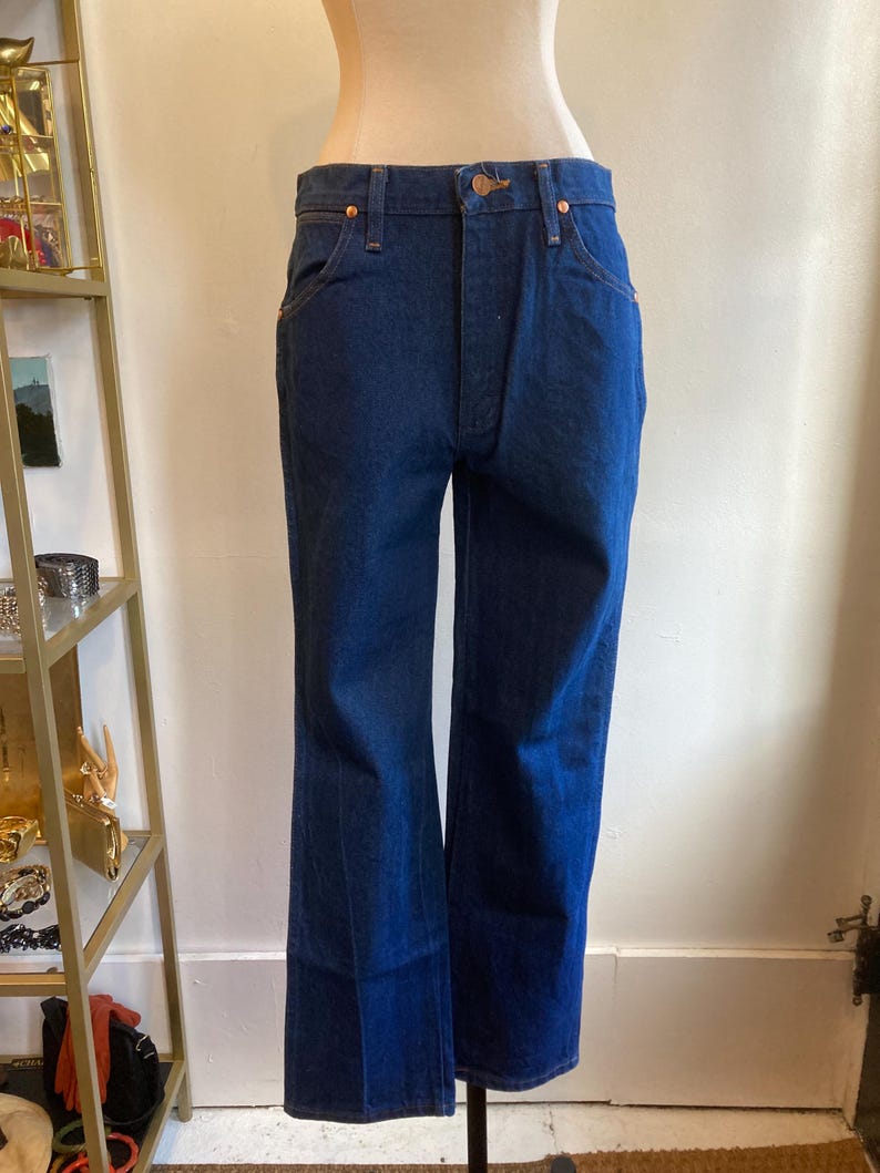 May include: Dark blue denim jeans with a classic five-pocket design and a slight flare at the hem. The jeans are displayed on a mannequin, showcasing the fit and style. The jeans have copper-colored rivets and a button closure.