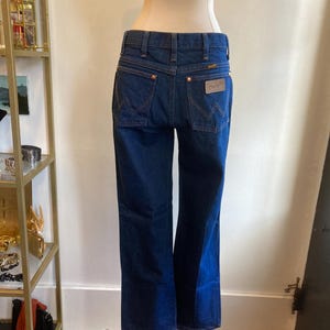 May include: Dark blue denim jeans with a classic five-pocket design. The jeans feature copper-colored rivets and a leather patch with the "Wrangler" logo on the back pocket. The jeans are displayed on a mannequin.