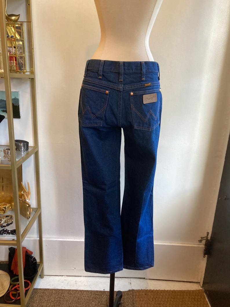 May include: Dark blue denim jeans with a classic five-pocket design. The jeans feature copper-colored rivets and a leather patch with the "Wrangler" logo on the back pocket. The jeans are displayed on a mannequin.