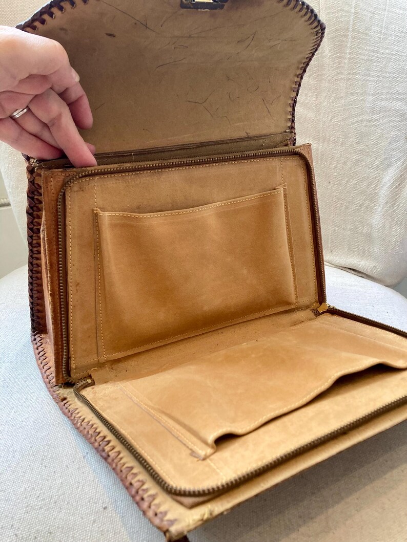 Pode incluir: Vista aberta de uma pasta de couro castanho claro vintage. A pasta tem fecho de correr, v&aacute;rios bolsos interiores e uma borda costurada decorativa. O interior &eacute; um tom mais claro do que o exterior.