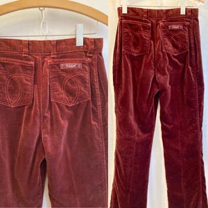 May include: A pair of burgundy velvet pants with a high waist and a wide leg. The pants have a back pocket with the word "Vidal" embroidered on it.