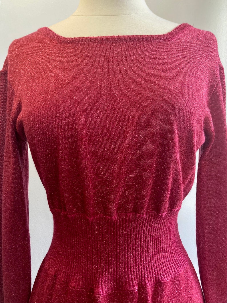 May include: A long-sleeved, burgundy knit dress with a fitted bodice and a flowing skirt. The dress has a subtle shimmer and a ribbed texture.