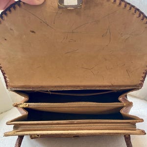 Pode incluir: Vista aberta de uma bolsa vintage de couro castanho claro com uma borda costurada. A bolsa tem um fecho de metal retangular e v&aacute;rios compartimentos com z&iacute;per. A al&ccedil;a &eacute; marrom mais escuro e o interior &eacute; um tom mais claro de marrom.