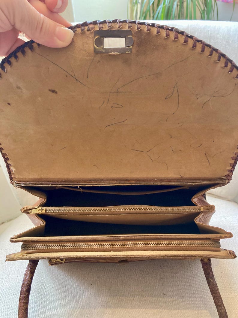 Pode incluir: Vista aberta de uma bolsa vintage de couro castanho claro com uma borda costurada. A bolsa tem um fecho de metal retangular e v&aacute;rios compartimentos com z&iacute;per. A al&ccedil;a &eacute; marrom mais escuro e o interior &eacute; um tom mais claro de marrom.