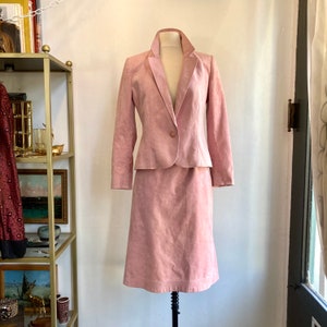 May include: A pink suede skirt suit with a blazer and a skirt. The blazer has a single button closure and a notched collar. The skirt is knee-length and has a straight silhouette.