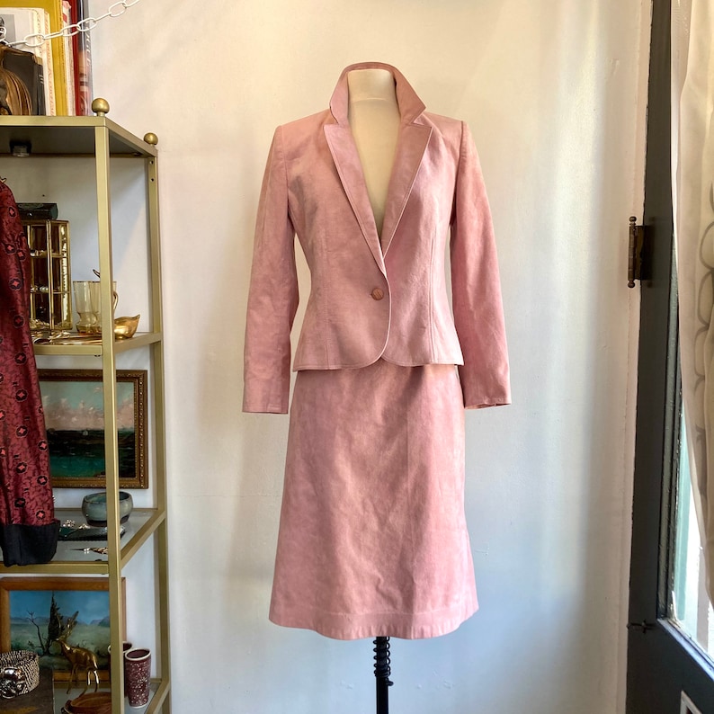 May include: A pink suede skirt suit with a blazer and a skirt. The blazer has a single button closure and a notched collar. The skirt is knee-length and has a straight silhouette.