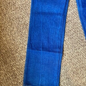 May include: Close-up of blue denim jeans. The jeans are a vibrant shade of blue and appear to be made of a sturdy denim material. The image shows the lower portion of the jeans, with the hem visible.