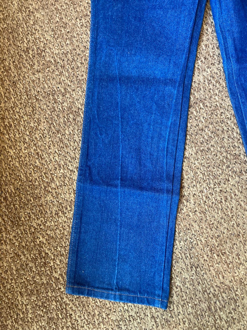 May include: Close-up of blue denim jeans. The jeans are a vibrant shade of blue and appear to be made of a sturdy denim material. The image shows the lower portion of the jeans, with the hem visible.