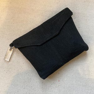Könnte beinhalten: Eine schwarze Clutch-Tasche mit Klappenverschluss. Die Tasche ist aus geripptem Stoff gefertigt und hat ein rechteckiges, klares Etikett am Reißverschlussanhänger. Die Tasche liegt auf einem neutralfarbenen Stoff.