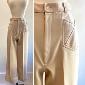 May include: Tan-colored wide-leg pants with a high waist and a button closure. The pants feature unique pocket designs with stitched detailing. The fabric appears to be a smooth, medium-weight material. The pants are displayed on a mannequin.