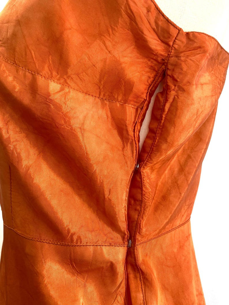 May include: A close-up of the back of an orange dress with a zipper closure. The dress is made of a shiny, silky fabric.