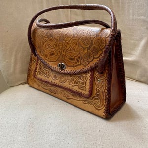 Pode incluir: Uma mala de m&atilde;o de couro castanho claro com um design floral em relevo. A mala tem uma pega superior, um fecho de aba e uma borda decorativa. O couro &eacute; de cor castanho claro com detalhes em castanho escuro.
