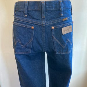 May include: Dark blue denim Wrangler jeans. The jeans have a classic five-pocket design with copper rivets and a leather patch on the back pocket. The stitching is a contrasting tan color, and the Wrangler logo is visible.