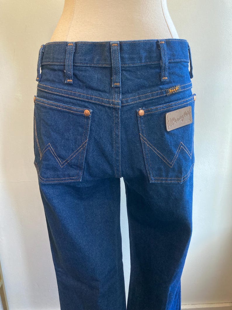 May include: Dark blue denim Wrangler jeans. The jeans have a classic five-pocket design with copper rivets and a leather patch on the back pocket. The stitching is a contrasting tan color, and the Wrangler logo is visible.