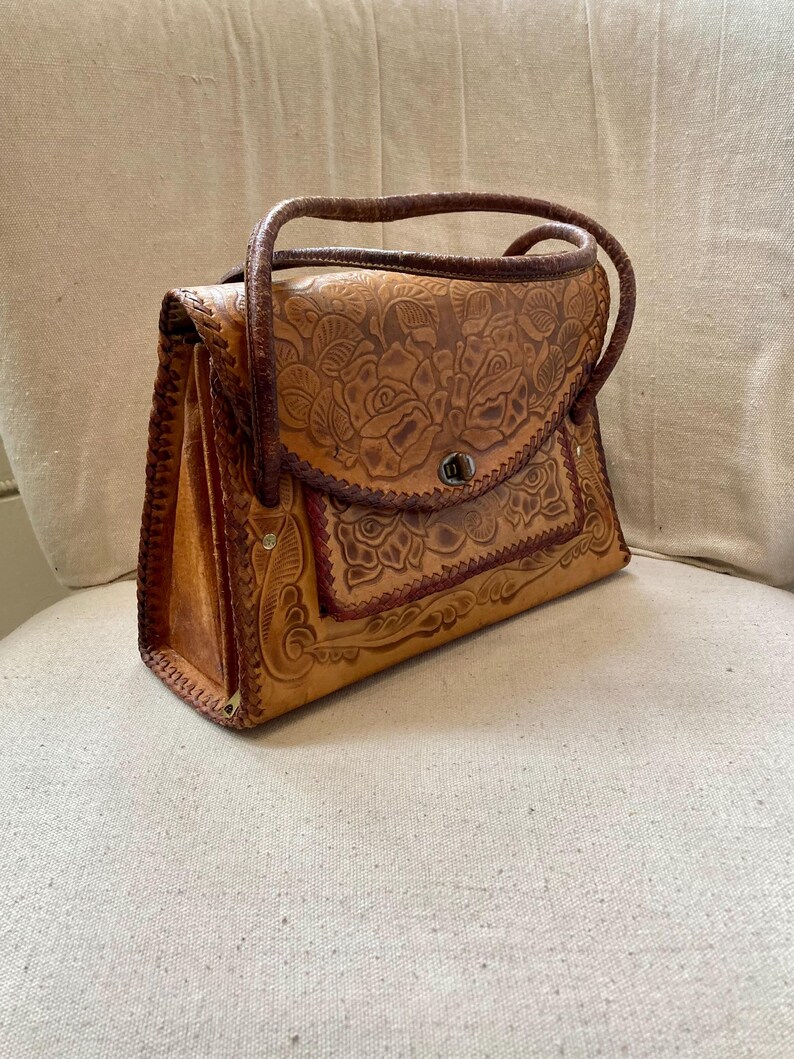 Pode incluir: Bolsa de m&atilde;o vintage em couro castanho com design floral em relevo. A bolsa apresenta uma al&ccedil;a superior, fecho de aba e costuras decorativas. A bolsa retangular parece estar em bom estado.