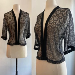 May include: Black lace bolero jacket with a scalloped pattern. The jacket has a V-neck, three-quarter sleeves, and black trim along the edges. The garment is displayed on a mannequin.
