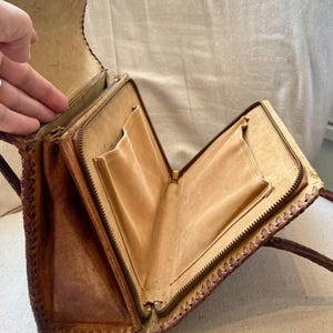 Pode incluir: Bolsa de m&atilde;o vintage de couro castanho com fecho de correr e v&aacute;rios bolsos internos. A bolsa apresenta um design de borda tran&ccedil;ada e uma al&ccedil;a longa. O interior &eacute; de cor bege claro e a bolsa parece desgastada.