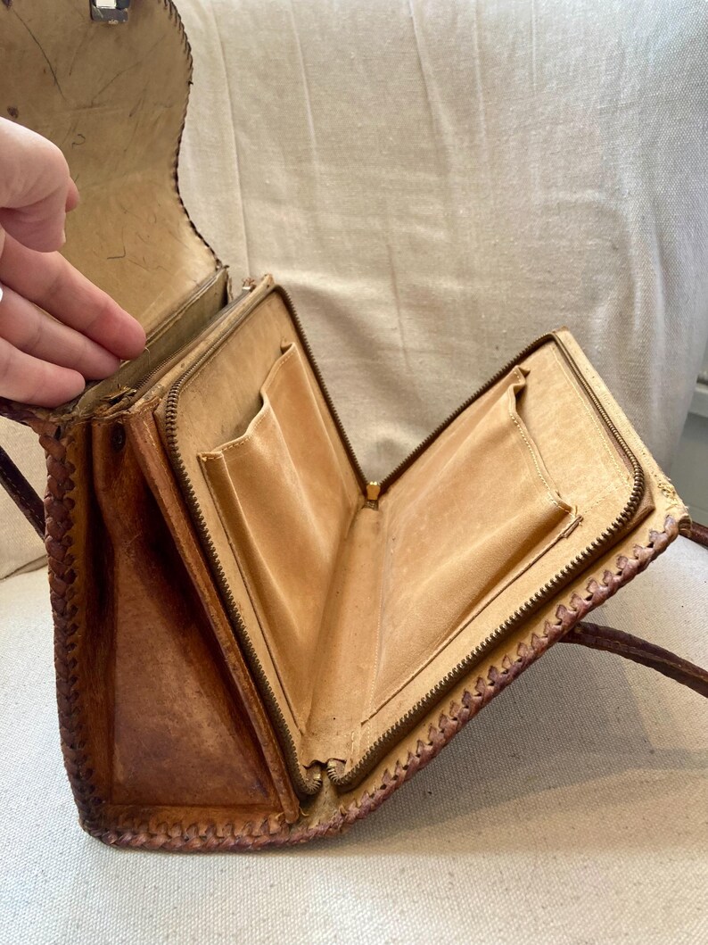 Pode incluir: Bolsa de m&atilde;o vintage de couro castanho com fecho de correr e v&aacute;rios bolsos internos. A bolsa apresenta um design de borda tran&ccedil;ada e uma al&ccedil;a longa. O interior &eacute; de cor bege claro e a bolsa parece desgastada.