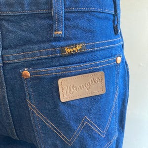 May include: Close-up of blue denim jeans with the Wrangler logo on the back pocket and waistband. The jeans have copper-colored rivets and stitching. The Wrangler logo is in a gold color.