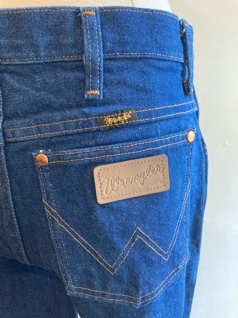 May include: Close-up of blue denim jeans with the Wrangler logo on the back pocket and waistband. The jeans have copper-colored rivets and stitching. The Wrangler logo is in a gold color.