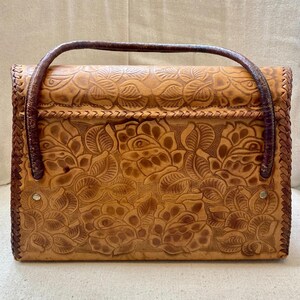 Pode incluir: Bolsa de m&atilde;o de couro castanho com um design floral em relevo. A bolsa apresenta uma al&ccedil;a e acabamentos em castanho escuro, com formato retangular e uma borda tran&ccedil;ada. O padr&atilde;o floral cobre a frente e as costas da bolsa.